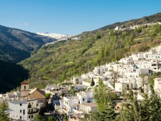 Pueblo de las Alpujarras con casas blancas en ladera, iglesia en primer plano y montañas al fondo bajo cielo azul.