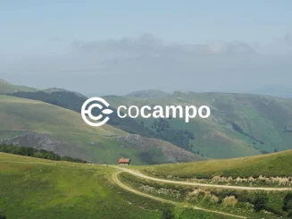 Paisaje rural de colinas verdes y montañas al fondo, con un camino de tierra que conduce a una pequeña casa; sobre la imagen aparece el logotipo de Cocampo.