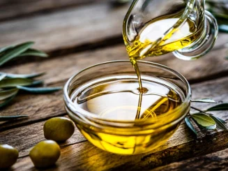 Aceite de oliva vertiéndose en un cuenco de cristal sobre una mesa de madera, con aceitunas y hojas alrededor.