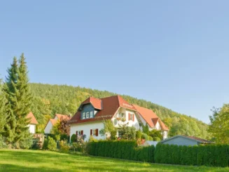 Casa unifamiliar blanca con tejado rojo, rodeada de jardín y setos, en una zona verde al pie de una ladera boscosa bajo un cielo despejado.