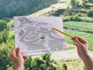 Una persona sostiene un plano de un terreno mientras señala una parcela específica en el mapa, con el fondo de un paisaje montañoso y tierras cultivadas.